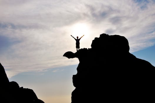Silhouette of a person celebrating on a mountain peak against a dramatic sky, symbolizing freedom and success.