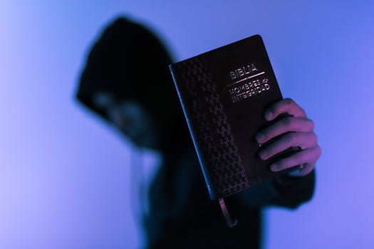 A hooded figure holds a Bible in low-light setting, emphasizing spirituality and mystery.