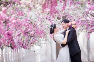 Bride and groom embrace under a pink floral archway in an outdoor wedding setting.